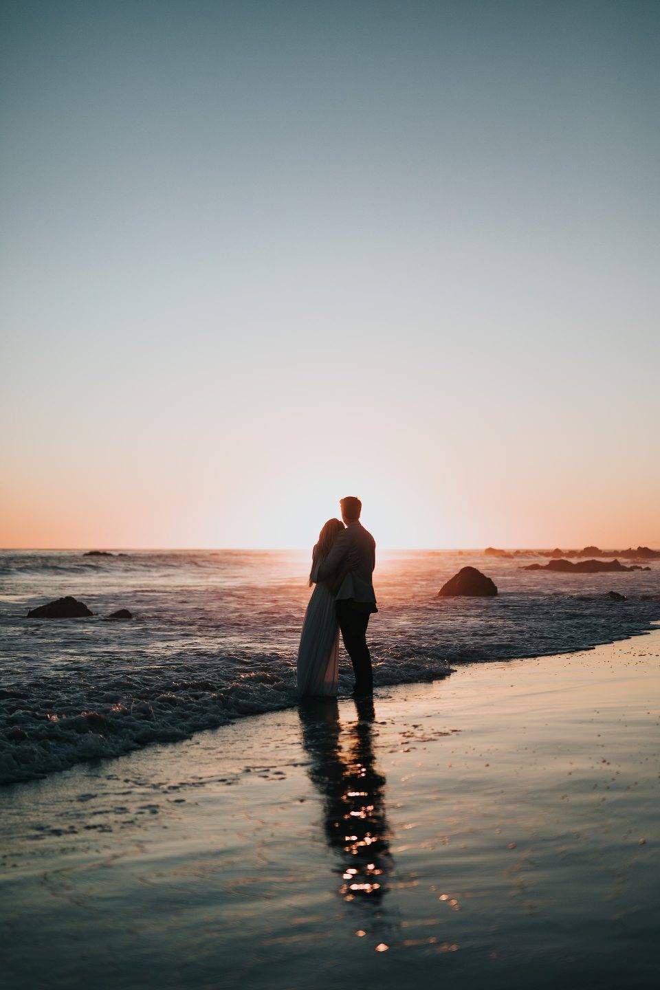 photo of couple standing on beach watching sunset